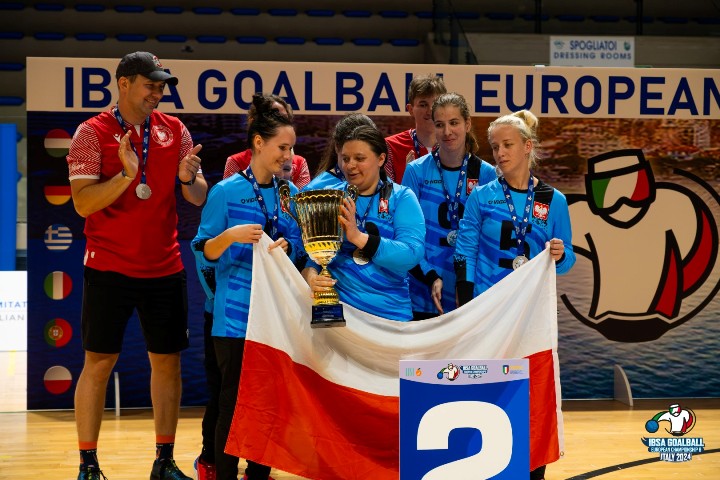 Reprezentantki Polski w goal ball. Cztery kobiety, w niebieskich bluzkach reprezentacji. Jedna z nich trzyma w ręku puchar. Pozostałe trzymają biało-czerwona flagę. 